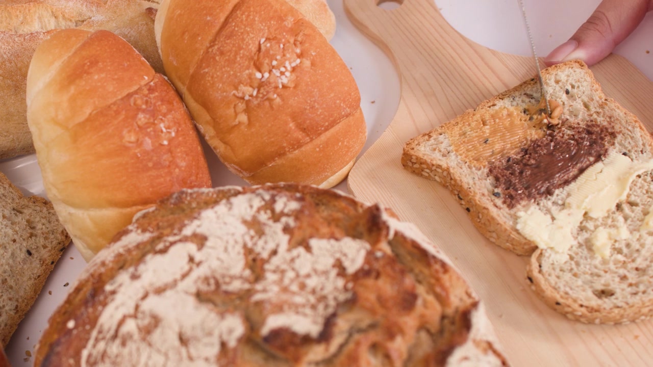 Close-up of a knife spreading peanut butter on toast beside sourdough, croissants, and baguettes.