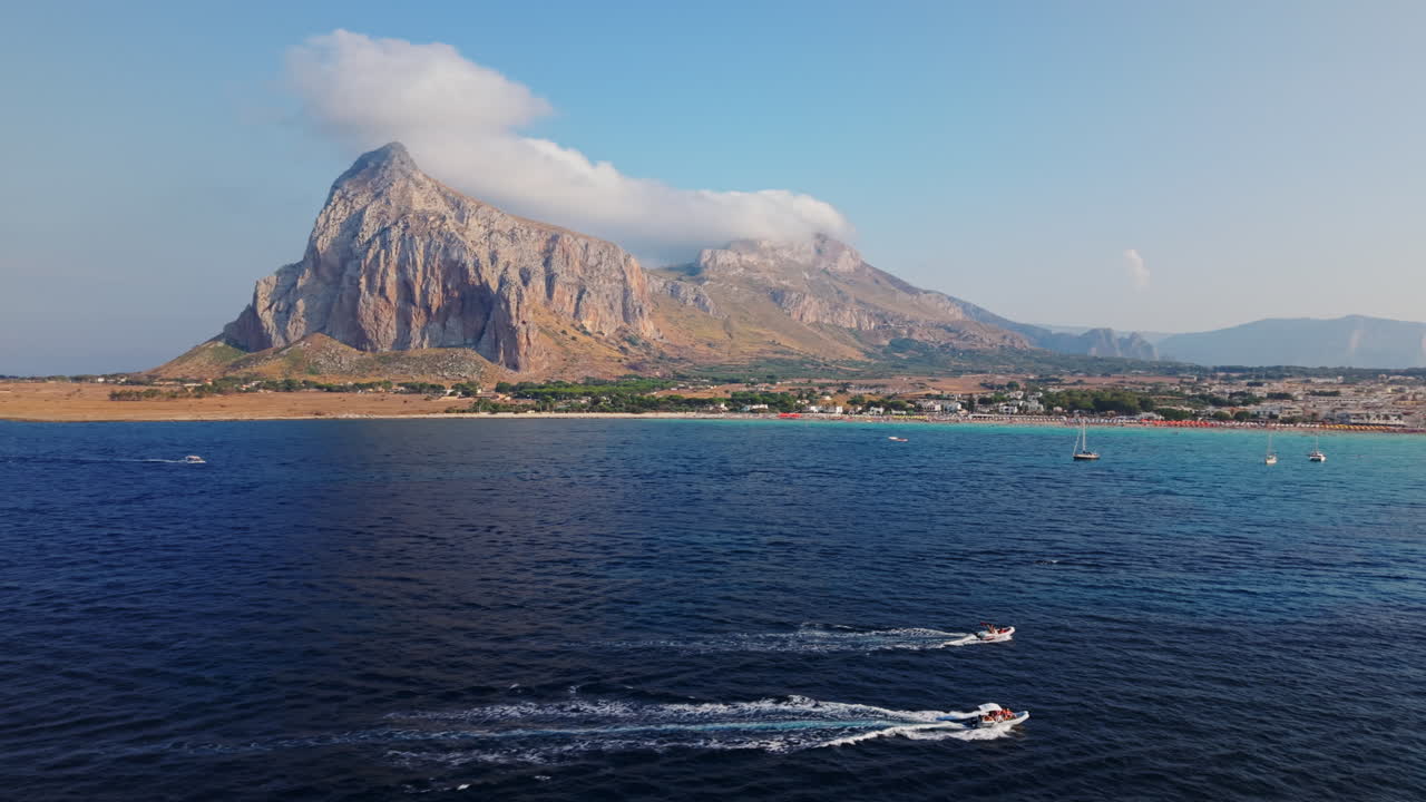 Drone moves toward Sicilian coast above speedboats, with rocky mountain ahead