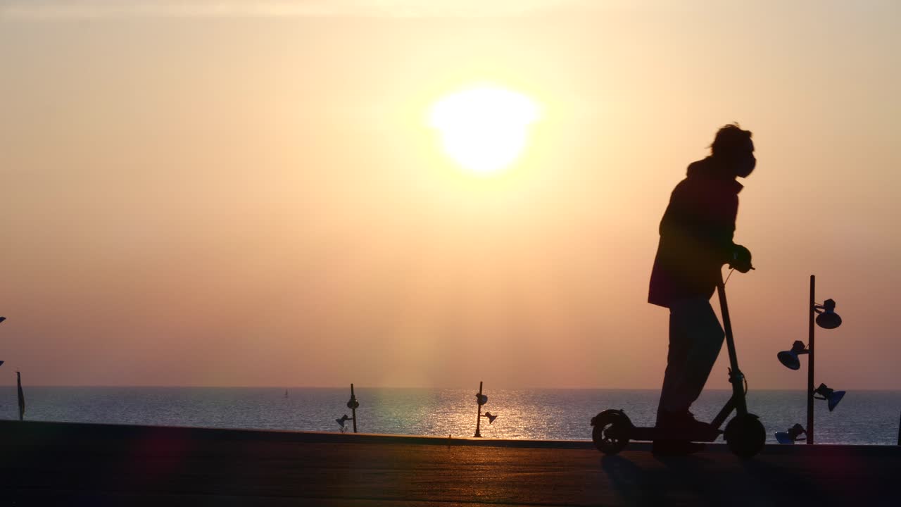 젊은 실루엣 남자는 해가 뜨면 해변에서 e스쿠터를 타고 경치를 감상하고 빠져나간다