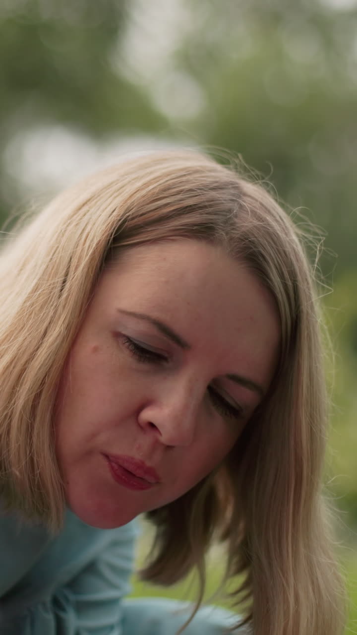 una mujer con un vestido azul claro parece estar soplando aire en algo atentamente, una vista parcial borrosa de alguien cerca de ella también es visible en primer plano