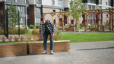 young male artist walks toward wooden bench in bright outdoor plaza with greenery and office building in background sits down and removes item from pocket while plants sway gently in breeze