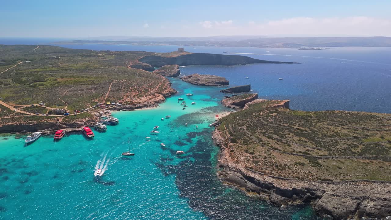 Lagoa Azul, Comino, Malta, e a vizinha Cominotto são mostradas de cima com águas turquesa, costa rochosa, barcos ancorados e paisagem mediterrânea circundante em um dia ensolarado de primavera