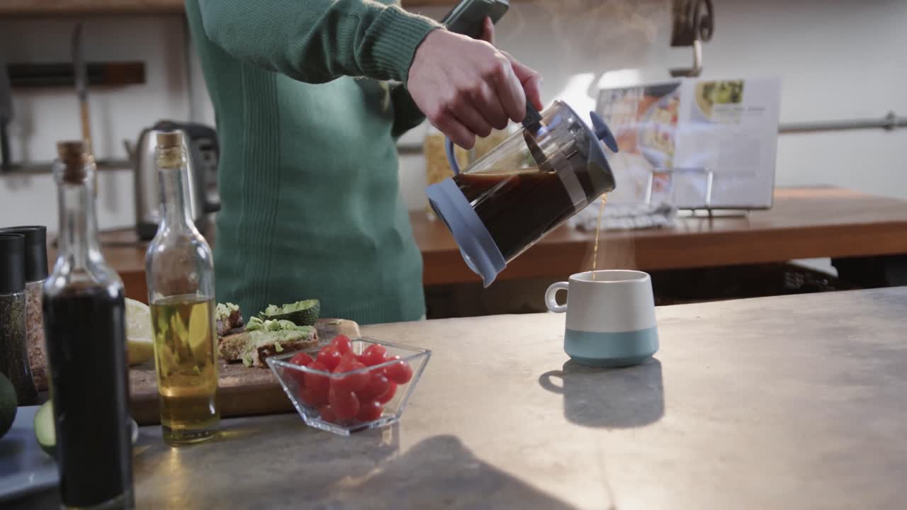 Mezzo busto di uomo caucasico che prepara il caffè e usa lo smartphone in cucina, al rallentatore.