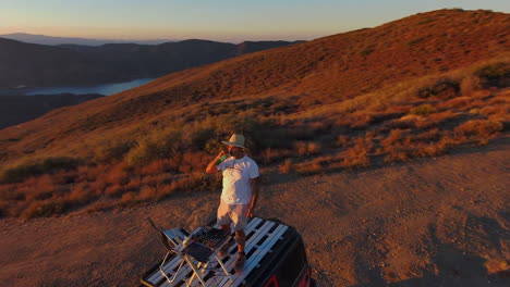 Man-danceing-alone-drinking-on-top-of-jeep-in-remote-desert-at-sunset,-aerial