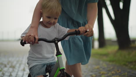 a mother guides her young child as he learns to ride a bicycle along a leafy pathway, the boy grips the handlebars while the mother holds his hand, supporting him as he moves forward