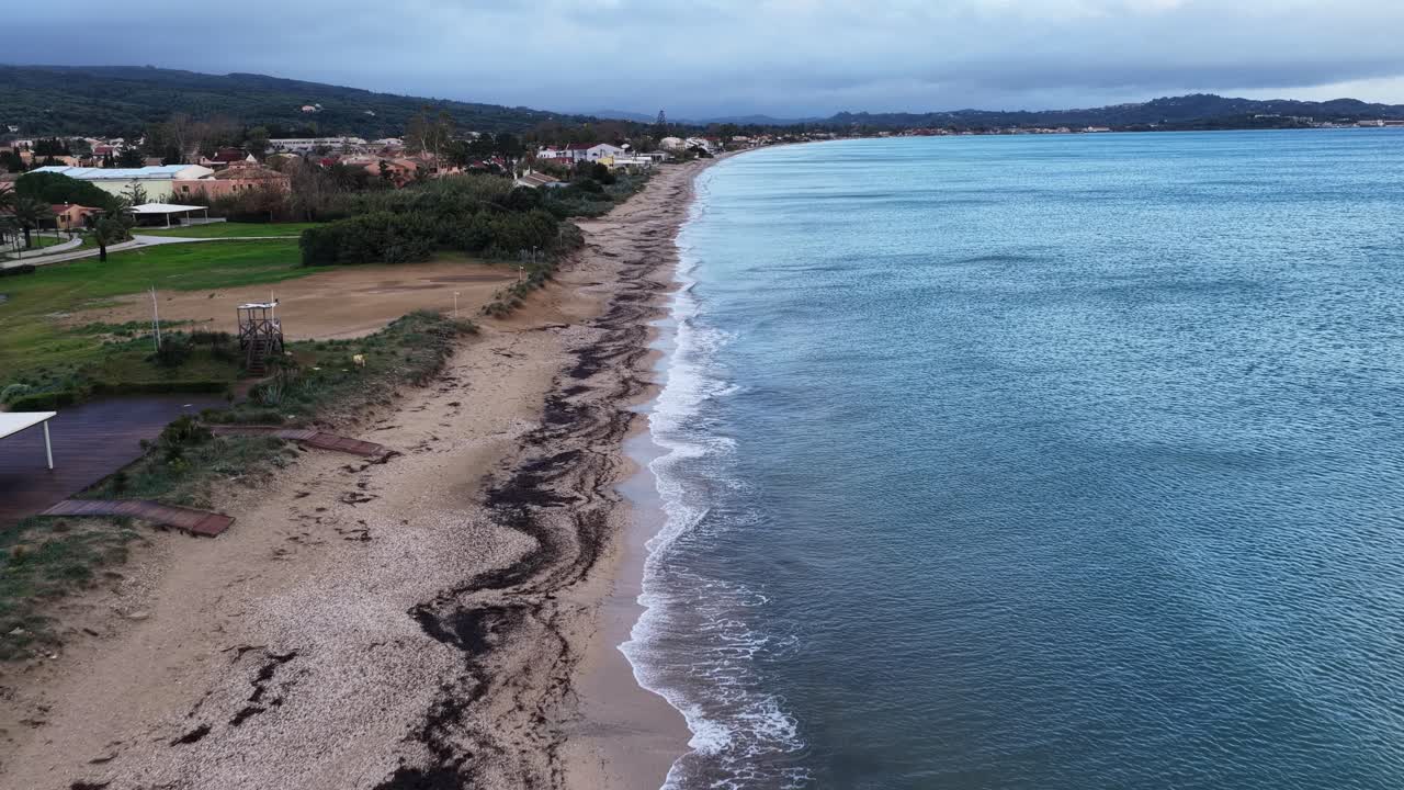 Long Sandy Coastline Aerial: Moody Sunrise or Sunset Drone View Over Tranquil Ocean Waves Breaking on Wide Beach. Mediterranean Seascape Panorama with Coastal Village in Distance and Overcast Sky