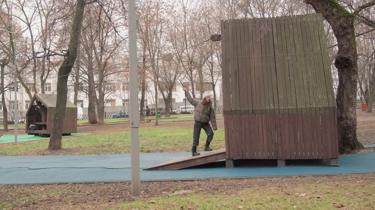 Mannlig skateboarder ruller over blått sikkerhetsunderlag på urban lekeplass, uformell jakke og jeans, dynamisk bevegelse forbi trelekeapparat, falne blader under hjulene, sent høstlys, ærlig.
