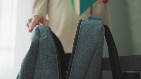 girl in casual beige sweater packing green notebook into blue school bag near window, preparing for class in soft morning light, organized school supplies visible, focused hand gesture emphasizes routine