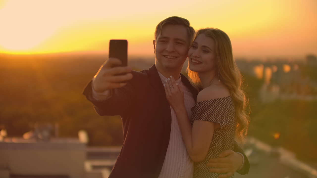 de pie en el techo al atardecer una pareja casada un hombre y una mujer se abrazan y se toman una selfie en el teléfono. toma fotos de ti mismo de pie en el tejado y abrazarse. jóvenes enamorados
