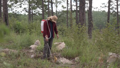 Long-shot-of-a-man-forest-ranger-working-with-a-backpack-sprayer-in-the-forest,-he-wears-a-red-shirt-and-a-hat