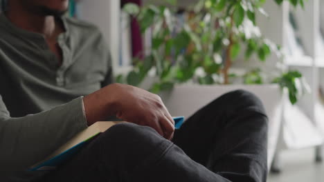 man with book sits on floor at home. smiling black guy enjoys reading interesting novel in comfortable atmosphere. student with textbook does homework