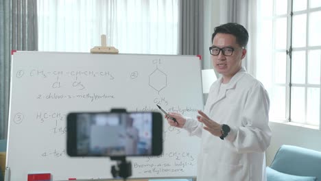 asian male teacher with glasses shoots video by smartphone camera while teaching chemist at home