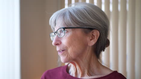 An-old-caucasian-woman-with-aging-face-gray-hair-and-glasses-staring-out-window-at-home-looking-hopeful,-calm-and-happy