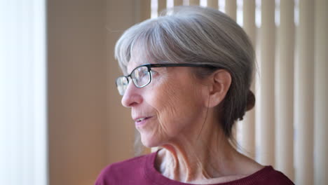 an old caucasian woman with aging face gray hair and glasses staring out window at home looking hopeful, calm and happy