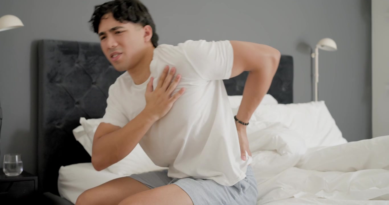 Man waking and sitting on bed, reacting to sudden torso ache, rubbing chest near glass