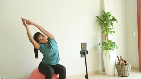 Ethnic-sportswoman-exercising-on-Pilates-ball-against-smartphone