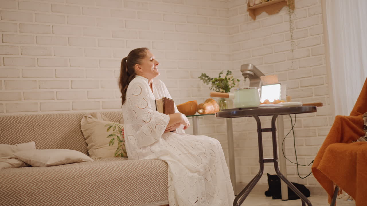 Relaxed couch tasting pumpkin pie while smiling and offering a slice, white woman as host and content creator enjoys warm autumn light and candle glow scene shows pumpkins, table lamp, vintage mixer