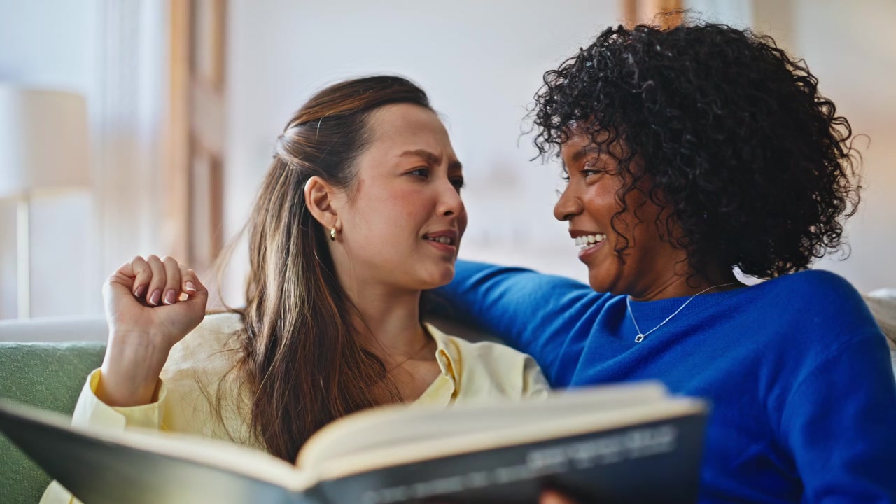 Friends reading book together sofa. Closeup multiethnic women laughing home