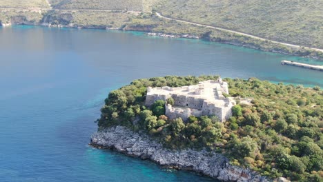 Vista-De-Drones-En-Albania-Volando-Lejos-De-Una-Isla-Verde-Con-Un-Fuerte-Medieval-Sobre-Agua-Azul-Clara-Del-Océano-En-Un-Día-Soleado