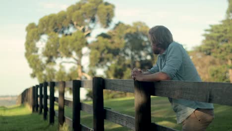 a young farmer in a blue shirt rests on a wooden fence post and looks out across the country