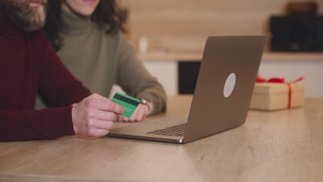 pareja comprando en línea con una tarjeta de crédito usando una computadora portátil sentada en una mesa cerca de un regalo