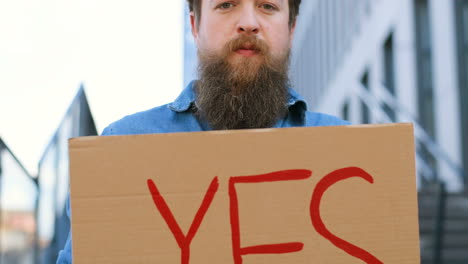 close-up view of bearded caucasian man holding yes" signboard and looking at camera in the street"