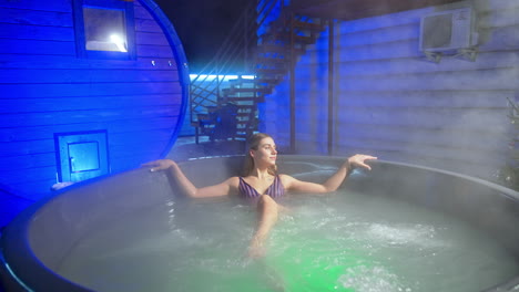 beautiful caucasian woman sits in the hot tub outdoors. lady is relaxing and lifts her arms posing for camera.