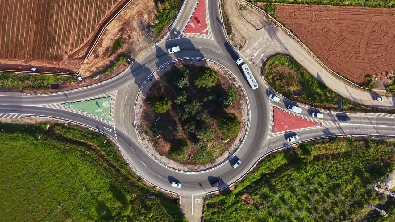 Cars drive around a roundabout with green and red lanes in a rural area near farmland on a sunny day