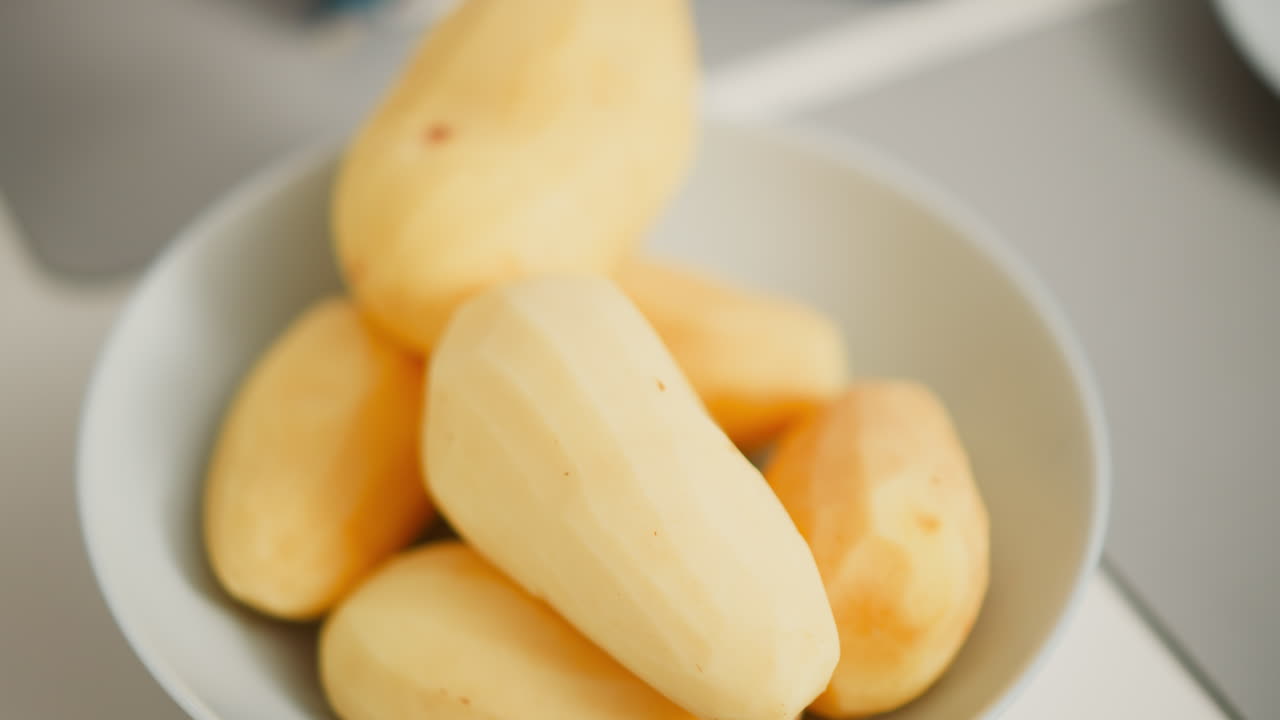 Close up of peeled potatoes stacked in bowl on white surface with person adding another freshly peeled potato, natural daylight softly illuminating smooth yellow surface and kitchen in background