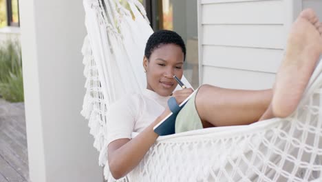 happy african american woman lying in hammock on terrace, writing in notebook, slow motion