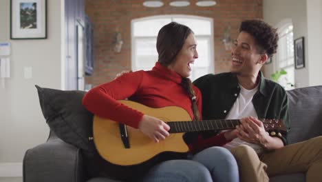 Happy-biracial-couple-sitting-on-sofa-in-living-room-playing-guitar