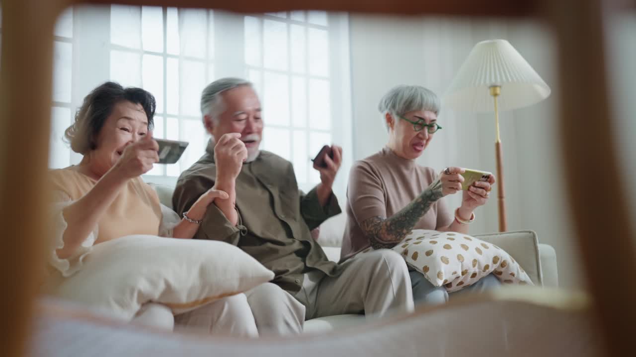happiness asian retired friend enjoy playing and competition game smartphone mobile online together on sofa in living room home interior background,asian group playing game together home isolate idea