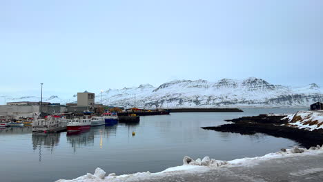 Waterfront-Panning-Coast-View-of-Djúpivogur-Small-Coastal-Village,-Fishing-Port-Harbor-in-Winter-Season,-Boats-and-Trawlers-Anchored-in-Water,-Snowy-Mountains-in-Background