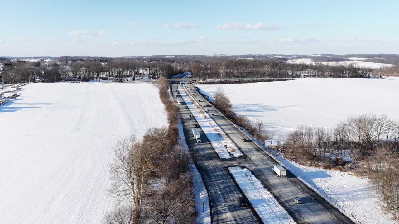 Traffico invernale su un'autostrada rurale vicino al confine tra Pennsylvania e Maryland, con auto che si muovono attraverso campi innevati sotto cieli limpidi e la bassa luce solare invernale. Paesaggio rurale. Ampia ripresa aerea dal basso verso l'alto