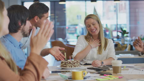 Colleagues-Celebrating-Businesswoman's-Birthday-At-Meeting-Around-Table-In-Modern-Open-Plan-Office