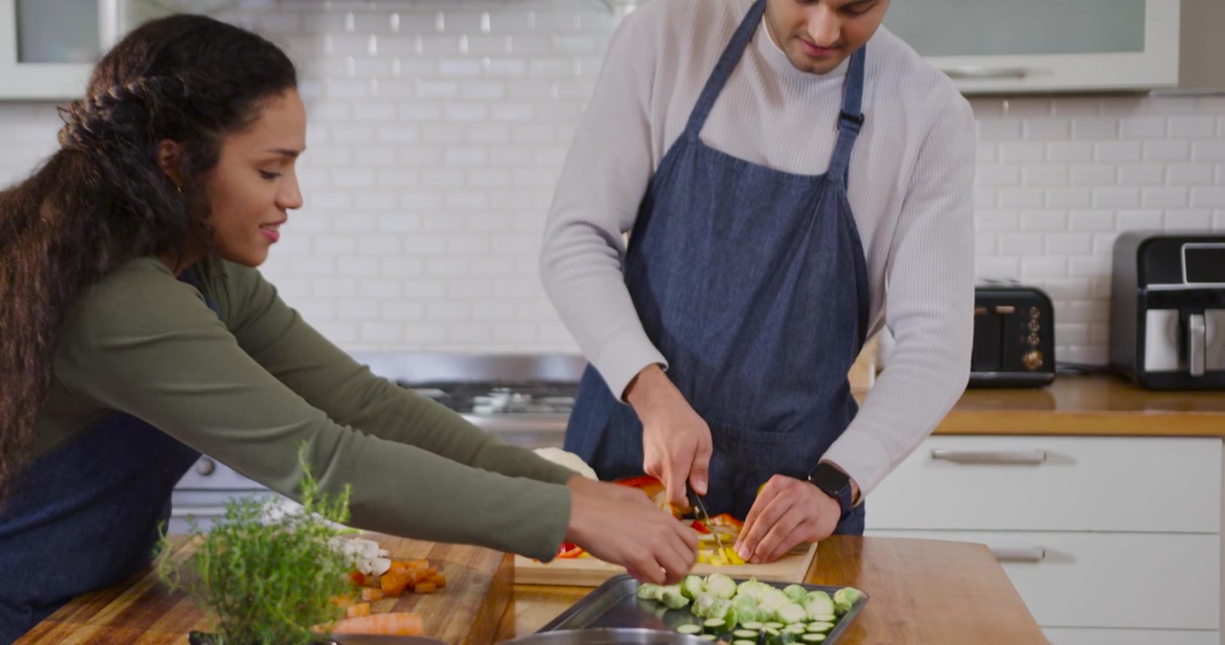 Pareja afroamericana con delantales picando, rebanando y colocando verduras en una isla de madera, preparando una comida.