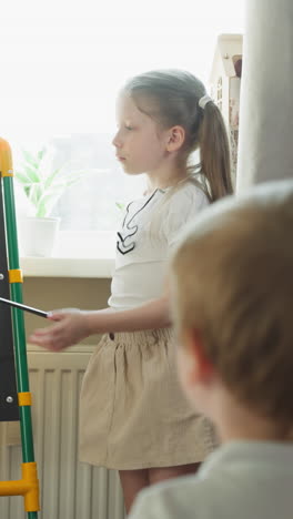 serious little girl shows maths tasks on blackboard to brother at home. sister teaches mathematics with junior boy in children room. playing school