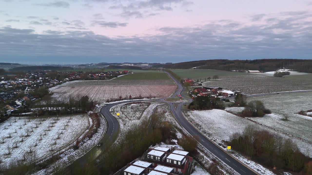 Aerial flyover winter landscape in small town with roundabout and hills in distance. Driving car on road in suburb neighborhood. Wide shot.
