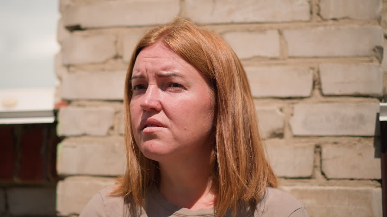 Donna tesa con capelli castano chiaro appoggiata a un muro di mattoni sotto la forte luce del sole, che socchiude leggermente gli occhi mentre guarda di lato, espressione che mostra stress e fatica durante un tranquillo momento all'aperto