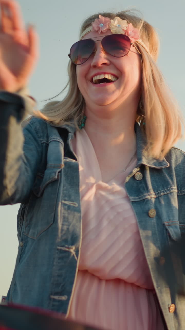 A cheerful woman wearing sunglasses and a flower headband is dancing with her hands raised and smiling while standing next to a red motorcycle in a grassy field