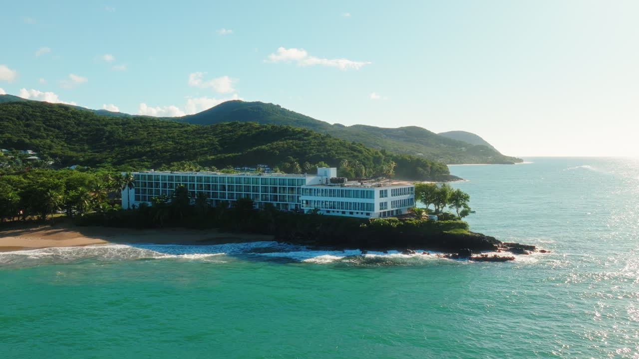 Aerial drone moves toward a large seaside hotel complex in Guadeloupe, revealing the coastline, palm trees, beach, and surrounding green hills