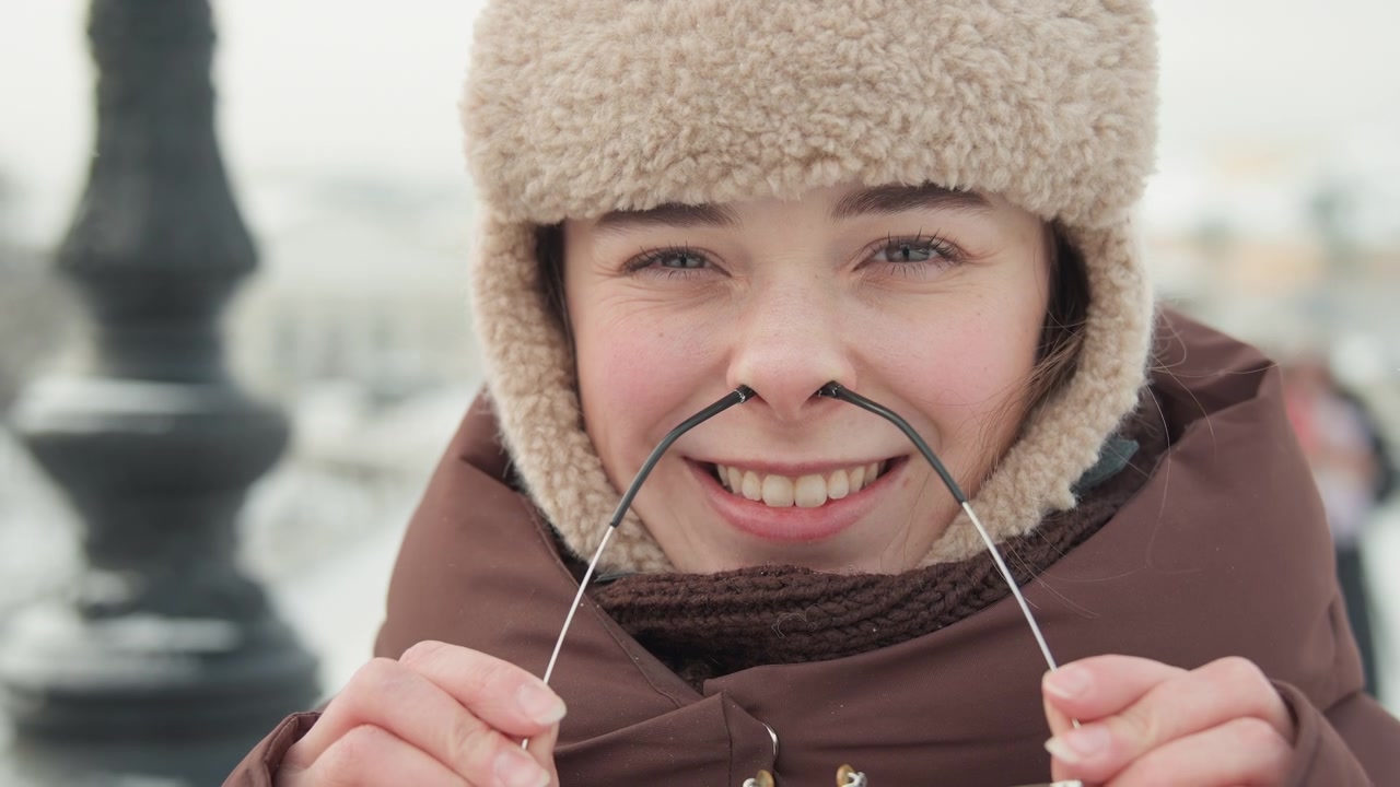Close-up eye-level portrait of young woman in snowy urban setting gently lowering metal eyeglass frames with both hands while smiling with relaxed, expression; wearing fluffy fur hat, padded