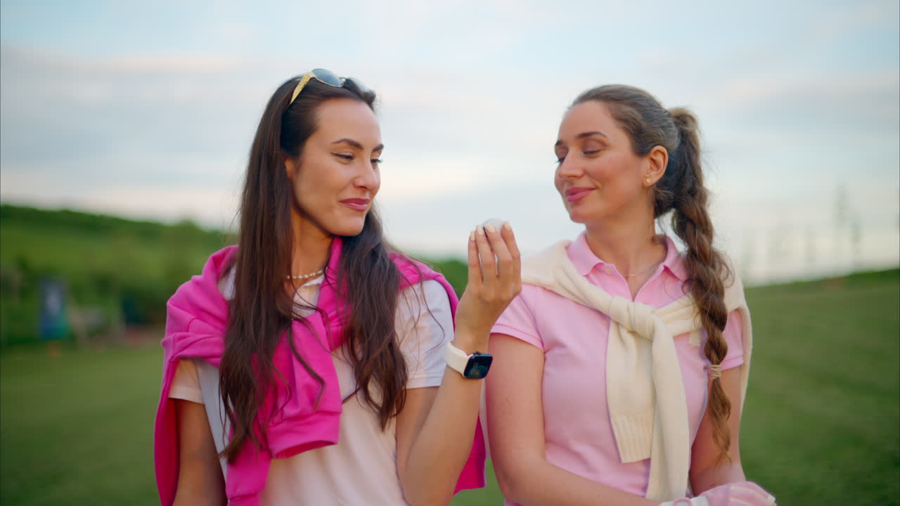 Two women in white and pink clothes holding a white golf ball on the course