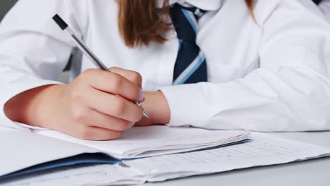 Close-Up-Of-Female-High-School-Students-Wearing-Uniform-Writing-In-Exercise-Book-At-Desk