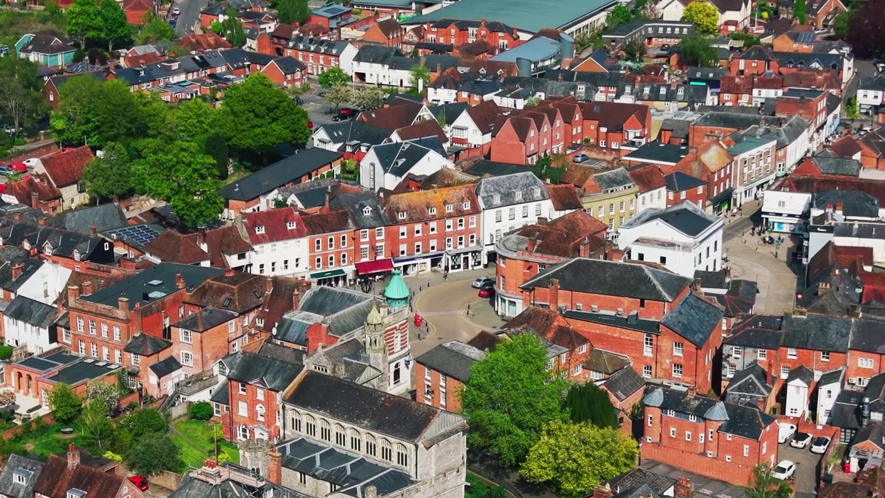 Bredt luftfoto fra drone af Romsey centrum i Hampshire, der viser historiske bygninger, hustage, gader, butikker, veje, træer, boligområder og byens overordnede layout på en solskinsdag.