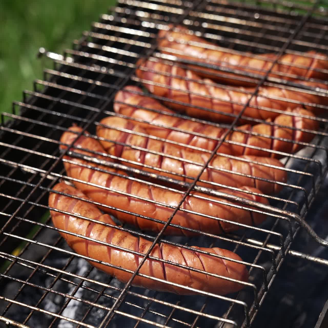 Roast wiener on a barbecue grill on charcoal. Cook turns over hot grilled sausages with a food tong in the garden in summer. Delicious food for a picnic