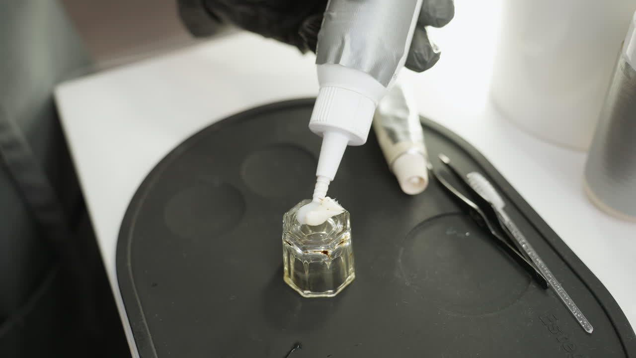 Close-up view of technician squeezing second cream into small transparent glass jar on black silicone tray with beauty tools like tweezers and applicator visible, showing preparation of cosmetic materials