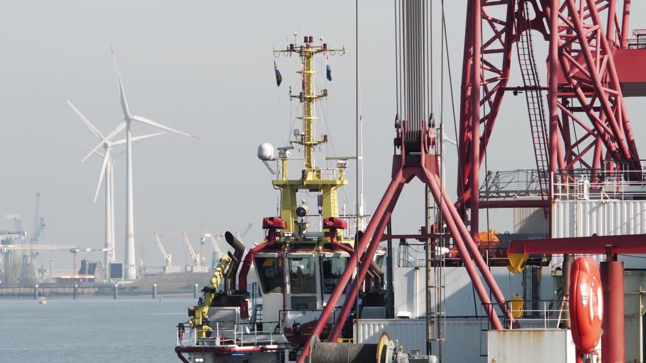 Close-up of a tugboat and red crane vessel in an industrial harbor, with wind turbines in the background