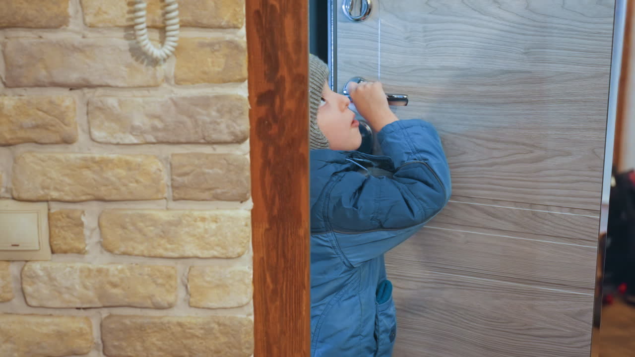 Young son peeking through apartment door, coat and knit hat next to brick wall, reaching for handle and intercom, curious expression, waiting for mom, soft indoor light, quiet hallway moment