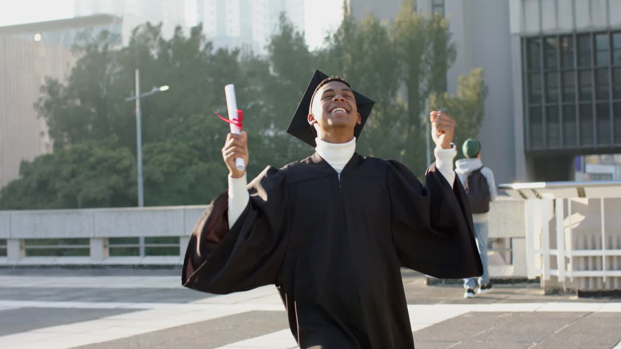 Male graduate in gown entering plaza, celebrating pumping fists, swinging diploma overhead laughing